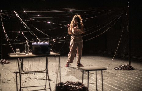 Photo 4 Une femme est seule sur scène, proche d'une table avec une radio, et des bandes magnétiques sont tirées.
