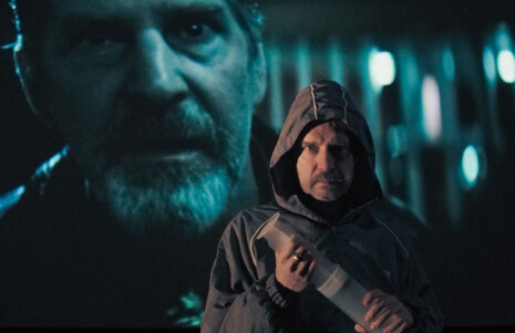 Photo 3 : Un homme sur scène, avec, derrière lui, un écran où est projeté un gros plan sur le visage d'un homme.