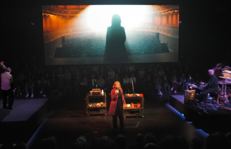 Photo 4 : Une femme est sur scène, avec des musiciens. Elle tient un micro et derrière elle se trouve un écran où est projeté un film.