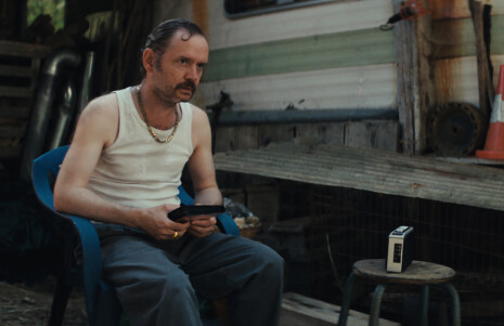 Photo 6 : Un homme est assis sur une chaise à l'extérieur, dans un cadre délabré. Il tient un objet dans sa main.